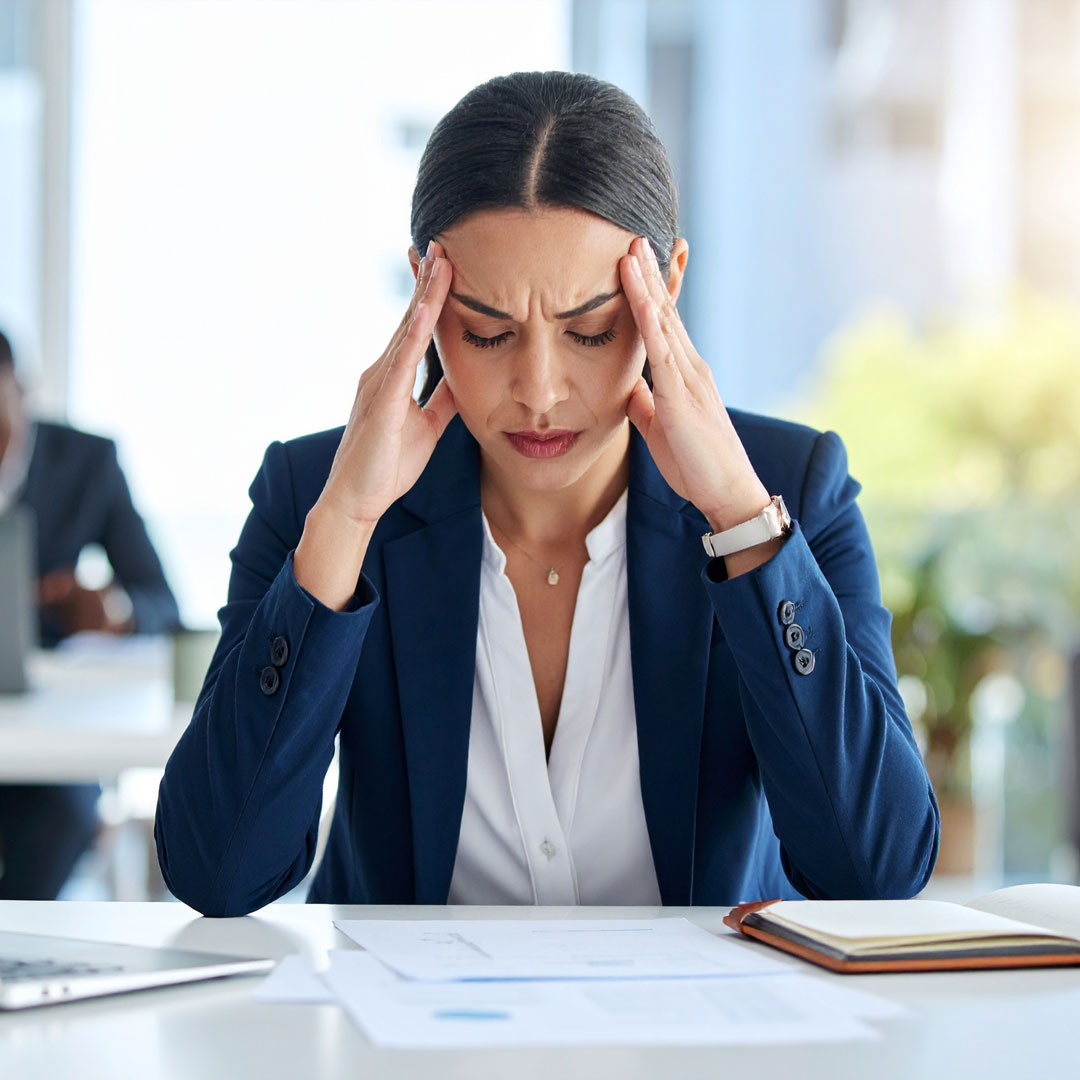 A woman sits at her desk in an open-plan office, her fingertips pressed to her temples and her expression revealing she is under stress